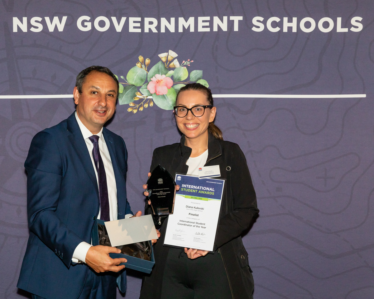 A man and woman stand in front of a purple screen, with the woman holiding a trophy and certificate